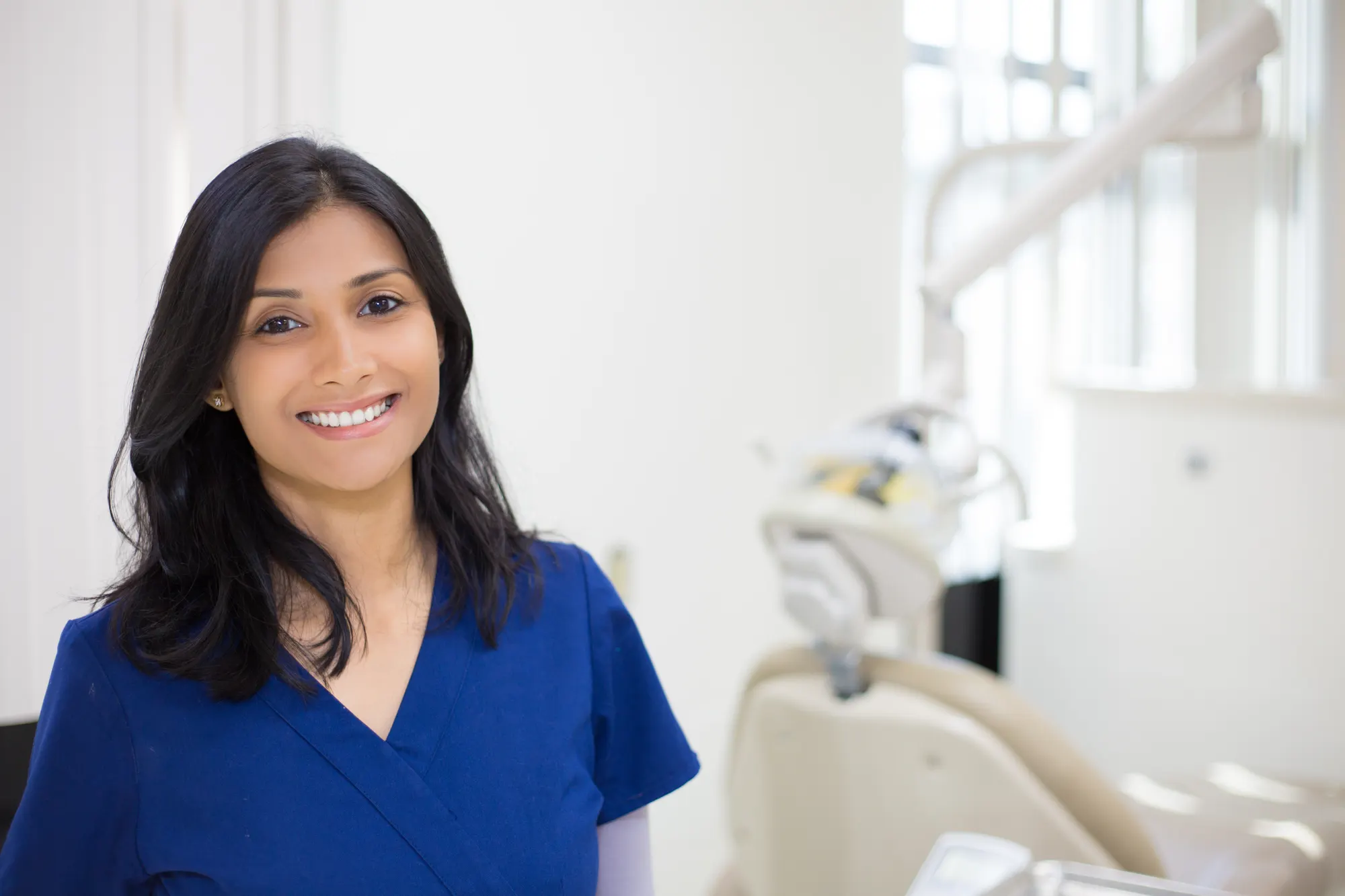 A smiling dental professional in blue scrubs in a treatment room
