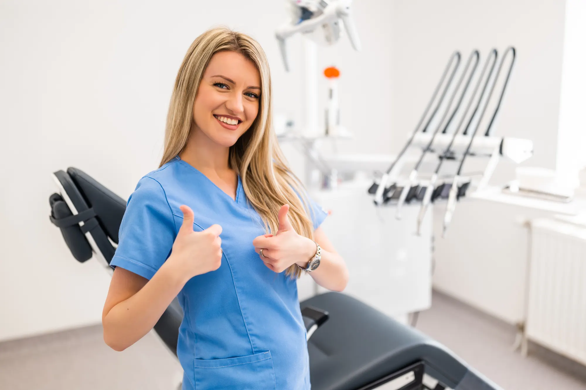 A smiling dental professional in blue scrubs giving a thumbs up beside a dental chair