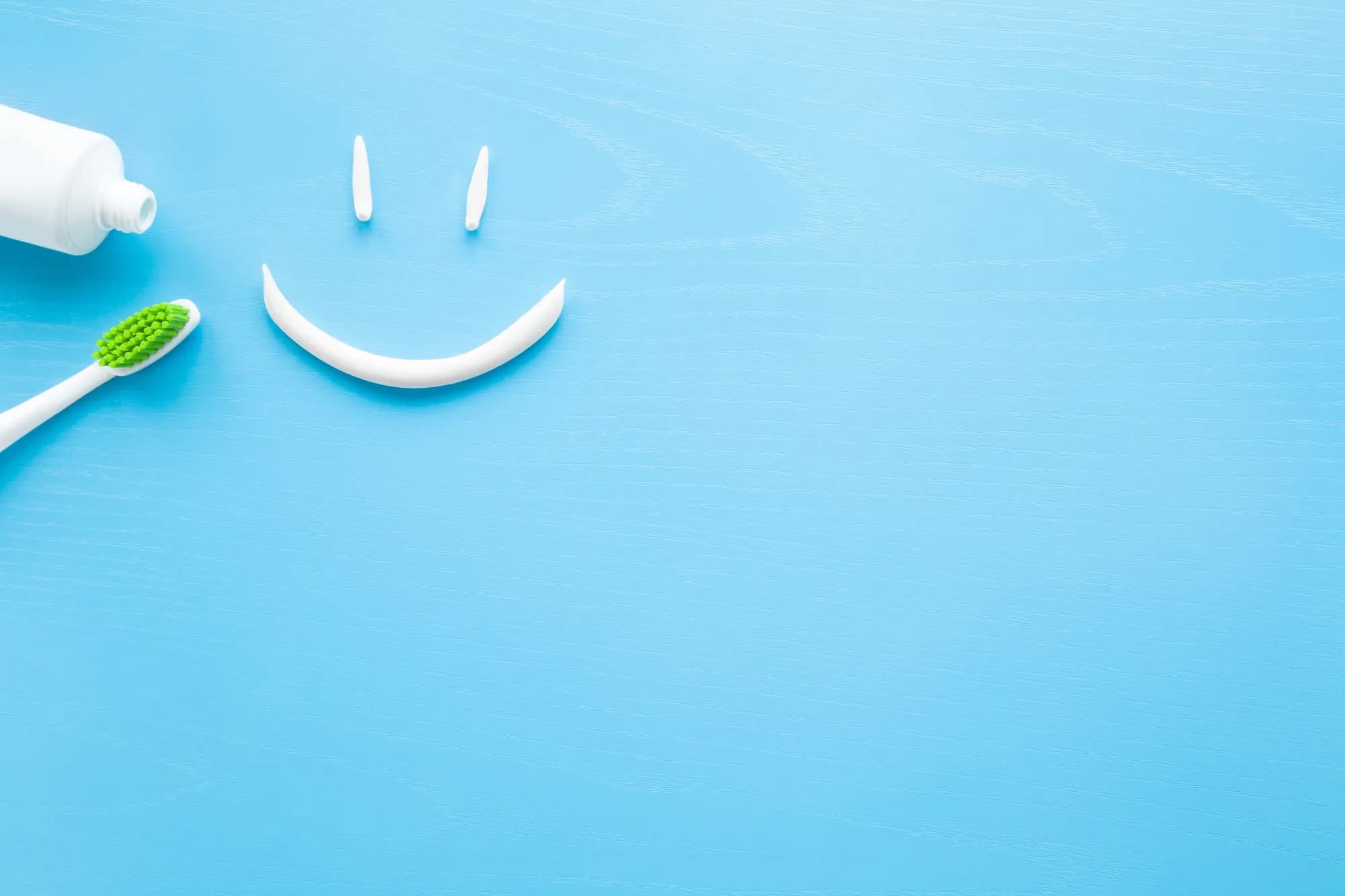 A green toothbrush and a tube of toothpaste with a smiley face drawn in paste, on a pale blue background